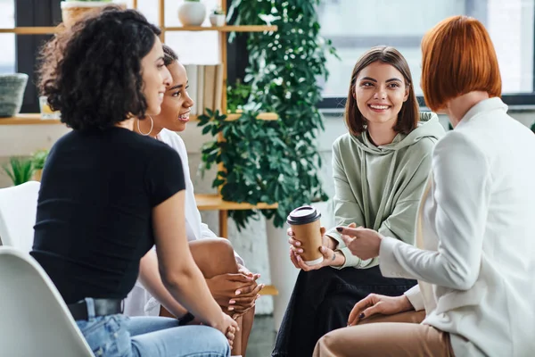 Happy young woman sitting with paper cup and listening to redhead psychologist during supportive therapy near multiracial girlfriends, understanding, support and mental health concept — Stock Photo