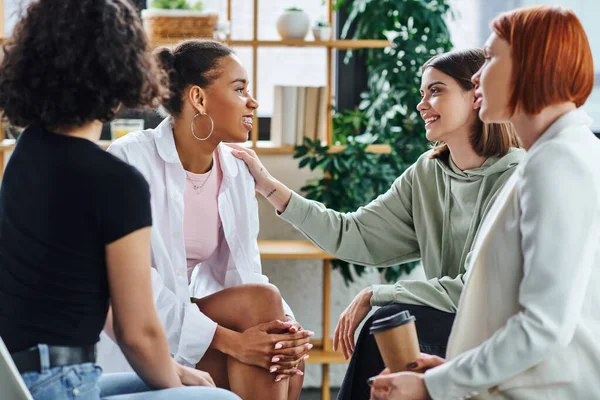 Young woman touching shoulder of pleased african american girlfriend while sitting next to redhead psychologist with paper cup during therapy in consulting room, mental wellness concept — Stock Photo