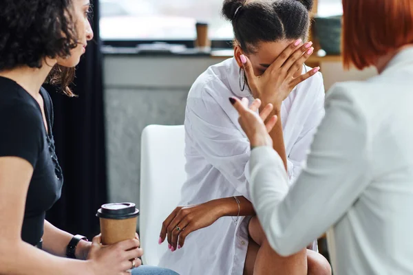 Depressed african american woman obscuring face with hand and crying near psychologist and multiracial friend sitting with coffee to go in consulting room, problem-solving concept — Stock Photo