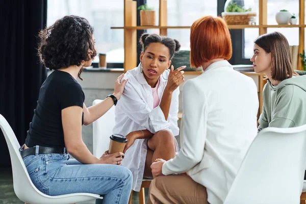 Woman with coffee to go touching shoulder of african american girlfriend and supporting her during psychology session in consulting room, problem-solving and mental wellness concept — Stock Photo