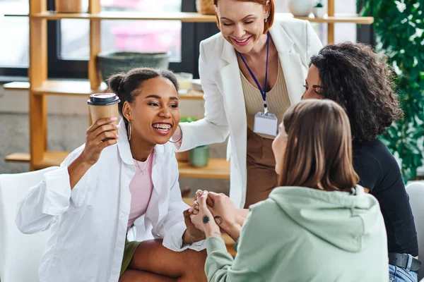 Multicultural women holding hands with carefree and joyful african american friend with coffee to go near smiling motivation coach in consulting room, problem-solving and mental wellness concept — Stock Photo