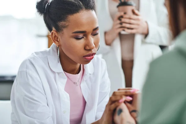 Young and thoughtful african american woman holding hands of girlfriend during supportive therapy near psychologist standing with paper cup on blurred background, empathy and problem-solving concept — Stock Photo