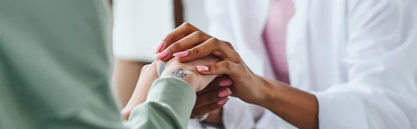 Partial view of african american woman holding hands of tattooed female friend while supporting and calming her during psychological therapy, empathy, solidarity and problem-solving concept, banner — Stock Photo