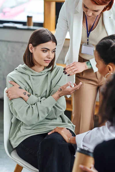 Mujer joven, triste y tatuada hablando con el entrenador de motivación mientras se sienta cerca de la novia afroamericana en primer plano borroso, empatía y concepto de resolución de problemas - foto de stock