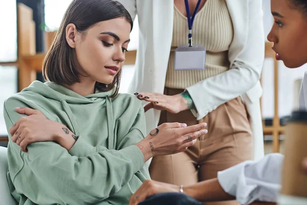 Professional psychologist calming disappointed tattooed woman sitting near african american female friend on blurred foreground in consulting room, empathy and problem-solving concept — Stock Photo