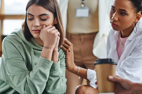 Young african american woman touching shoulder of frustrated tattooed girlfriend near motivation coach standing on blurred background in consulting room, empathy and female solidarity concept — Stock Photo