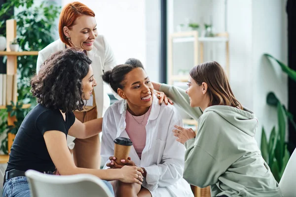Multicultural women embracing happy african american girlfriend near pleased redhead psychologist during group therapy in consulting room, friendship and mental wellness concept — Stock Photo