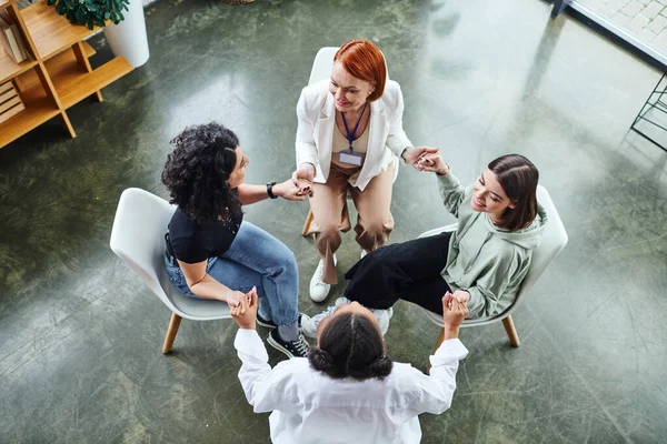 High angle view of joyful multiethnic female friends and redhead motivation coach sitting in circle and holding hands while talking on group therapy, communication and mental wellness concept — стоковое фото