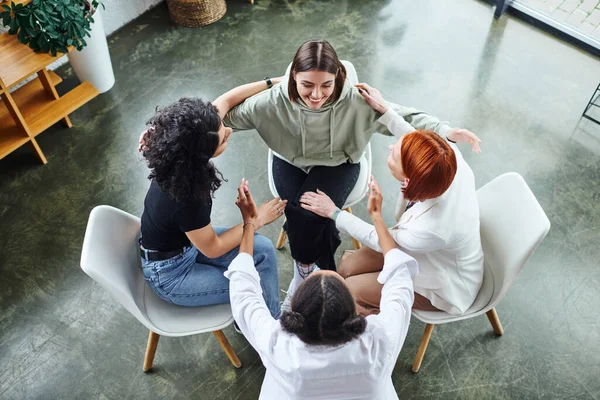 High angle view of motivation coach and optimistic multicultural female friends sitting in circle and embracing on group therapy in consulting room, communication and mental wellness concept — Stock Photo