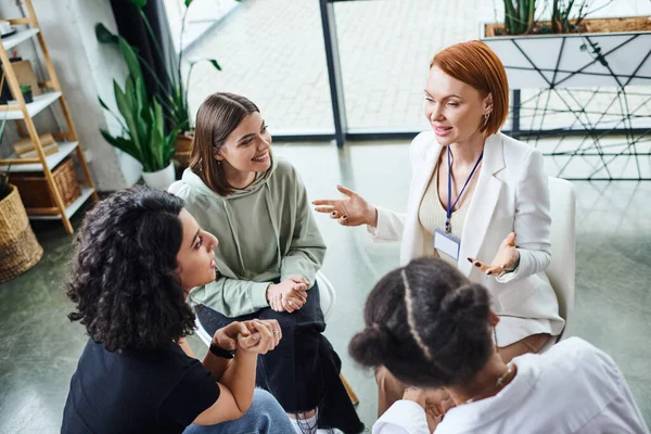 High angle view of redhead psychologist pointing with hands and talking to smiling multicultural women during motivation therapy in consulting room, friendship and mental wellness concept — Stock Photo