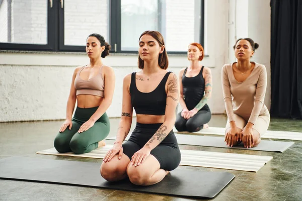 Groupe d'amies multiculturelles en vêtements de sport méditant les yeux fermés tout en pratiquant le yoga dans la pose de tonnerre, la paix intérieure et le concept de conscience du corps — Photo de stock