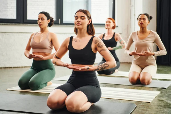Entrenador de yoga tatuado meditando con los ojos cerrados mientras está sentado en la pose del rayo cerca del diverso grupo de mujeres multiétnicas, paz interior y concepto de conciencia corporal - foto de stock