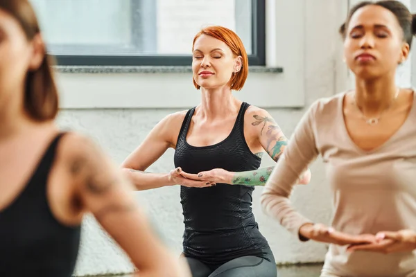 Tattooed and redhead woman practicing yoga and meditating with closed eyes near multicultural women on blurred foreground, inner peace and body awareness concept — Stock Photo