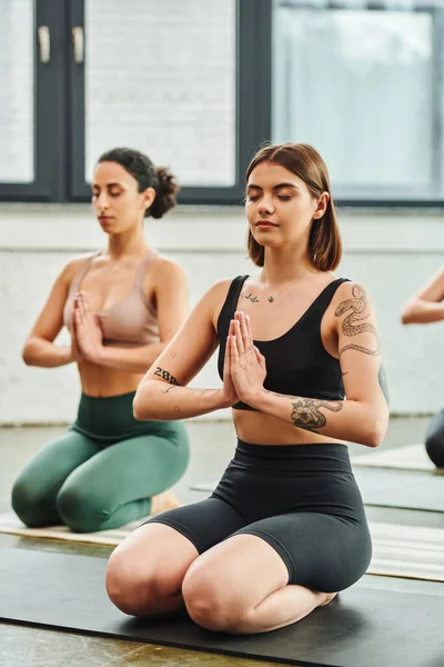 Young tattooed woman sitting in thunderbolt pose and meditating with closed eyes and praying hands near multiracial girlfriend on blurred background, inner peace and body awareness concept — Stock Photo