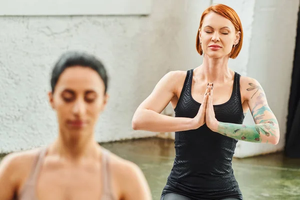 Tattooed and redhead woman in sportswear meditating with closed eyes and praying hands near multiracial female friend on blurred foreground, inner peace and body awareness concept — Stock Photo