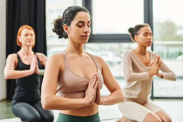 Multiracial woman in sportswear meditating with closed eyes and praying hands during yoga class near multiethnic girlfriends on blurred background, inner peace and body awareness concept — Stock Photo