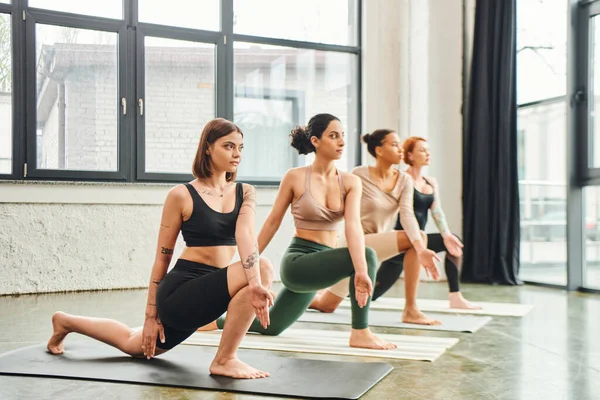 Longueur complète de copines multiculturelles dans les vêtements de sport pratiquant le yoga en fente sur la pose du genou et regardant vers l'avenir dans la salle de gym, harmonie et concept de santé mentale — Photo de stock