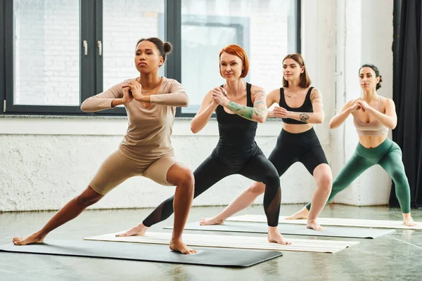 Full length of multiethnic female friends in sportswear standing in crescent lunge pose with clenched hands while meditating during yoga class, harmony and mental health concept — Stock Photo