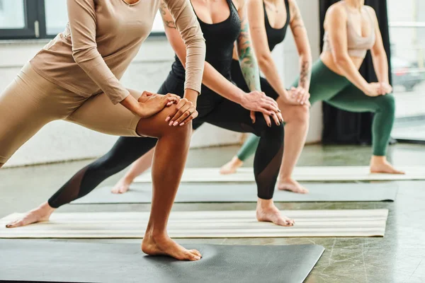 Cropped view of multicultural female friends in sportswear practicing yoga while standing in crescent lunge pose in gym, harmony and mental health concept — Stock Photo