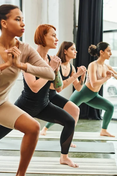 Diverse group of multicultural female friends in sportswear practicing yoga in crescent lunge pose with clenched hands, harmony and mental health concept — Stock Photo