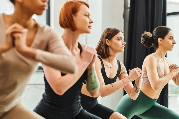 Junge und tätowierte Frau, die nach vorne blickt, während sie Yoga mit geballten Händen in der Nähe multiethnischer Freundinnen auf verschwommenem Vordergrund, Harmonie und psychische Gesundheit praktiziert — Stockfoto