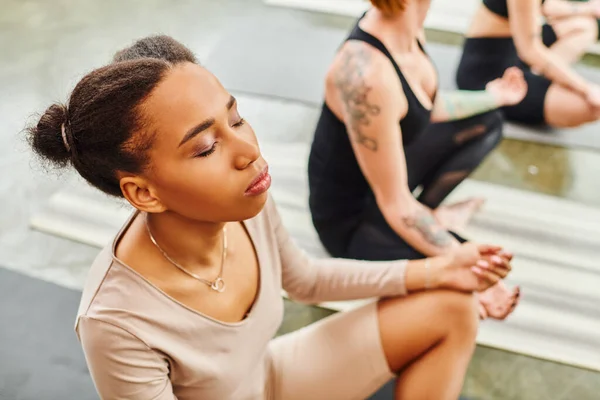 Vista de ángulo alto de la joven afroamericana con los ojos cerrados meditando durante la clase de yoga junto a amigas sentadas en pose fácil sobre fondo borroso, armonía y concepto de bienestar - foto de stock