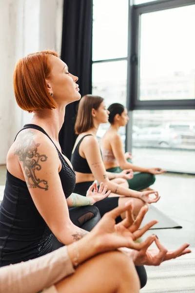 Ruiva tatuado mulher sentado em pose fácil com olhos fechados durante aula de ioga com grupo diversificado de amigos femininos multiétnicos, harmonia e conceito de bem-estar — Fotografia de Stock