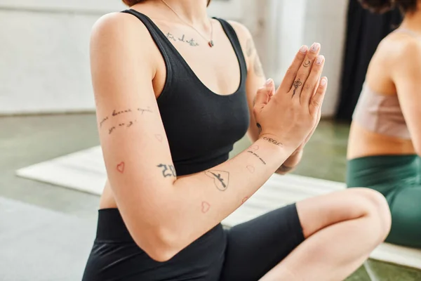 Обрезанный вид молодой татуированной женщины в спортивной одежде медитирующей в легкой позе с молитвенными руками, практикующей йогу рядом с другом в тренажерном зале, гармонии и оздоровительной концепции — стоковое фото