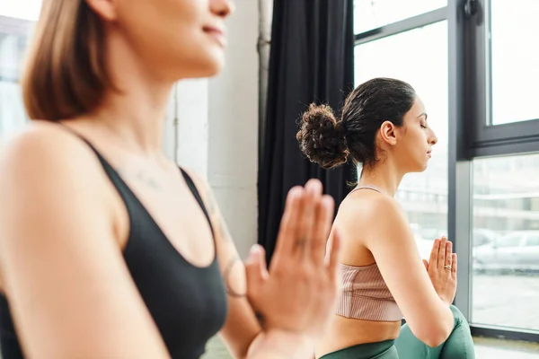 Multirassische Frau sitzt mit geschlossenen Augen und meditiert mit betenden Händen während eines Yoga-Kurses in der Nähe ihrer Freundin auf verschwommenem Vordergrund, Harmonie und Wellness-Konzept — Stockfoto