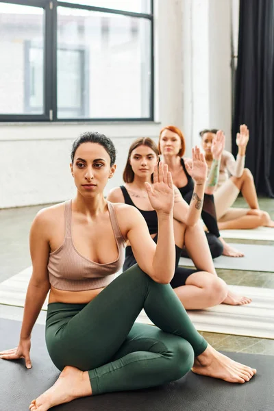 Diverse group of multiethnic female friends in sportswear meditating while sitting in lord of fishes pose and looking away during yoga class in gym, wellness and mental health concept — Stock Photo