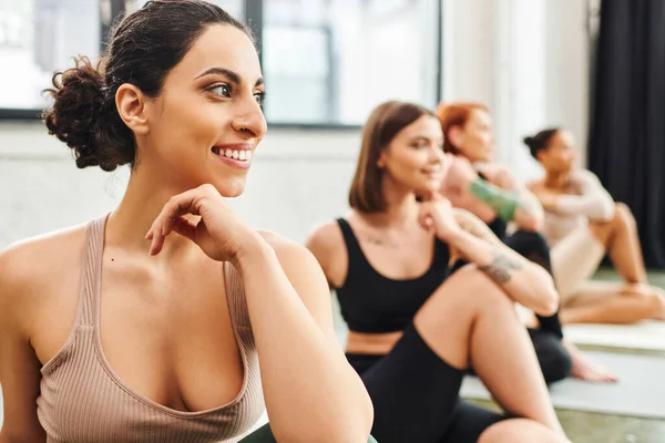 Mulher multirracial alegre olhando para longe durante a aula de ioga perto de grupo diversificado de namoradas multiétnicas em fundo turvo, bem-estar e conceito de saúde mental — Fotografia de Stock