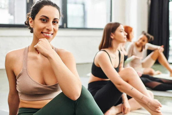 Fröhliche multiethnische Frau in Sportbekleidung, die in die Kamera schaut, neben multiethnischen Freundinnen, die auf verschwommenem Hintergrund während des Yoga-Kurses sitzen, Wellness und Konzept für psychische Gesundheit — Stockfoto