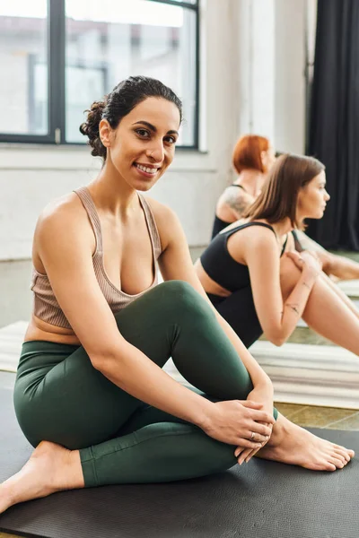 Joyful multiracial woman in sportswear sitting and smiling at camera next to female friends practicing yoga in gym on blurred background, wellness and mental health concept — Stock Photo