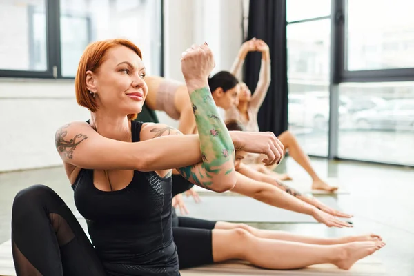 Happy, redhead and tattooed woman in sportswear training in gym while multiethnic female friends practicing yoga poses on blurred background, wellness and mental health concept — Stock Photo