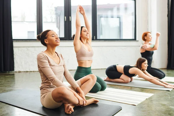 Joven y sonriente mujer afroamericana sentada en pose fácil y meditando con los ojos cerrados mientras amigos multiétnicos entrenan durante la clase de yoga, el bienestar y el concepto de salud mental - foto de stock