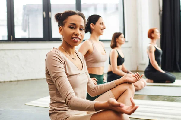 Positive and young african american woman with braces meditating in easy pose and looking at camera next to multicultural friends practicing yoga in gym, wellness and mental health concept — Stock Photo