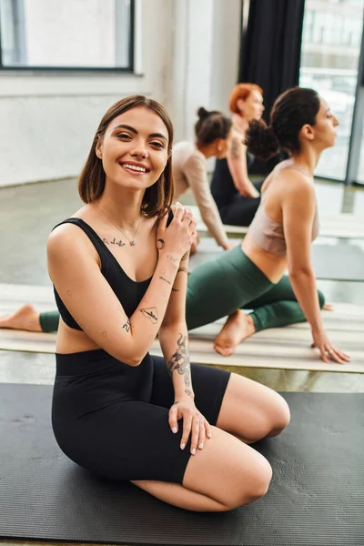 Молодая и счастливая татуированная женщина в спортивной одежде смотрит в камеру, сидя рядом с мультикультурными подругами, медитирующими на занятиях йогой, оздоровлении и психическом здоровье — стоковое фото