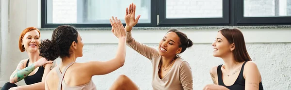 Jovem, despreocupado e animado afro-americano mulher dando alta cinco para amigo perto de mulheres alegres durante aula de ioga em ginásio, amizade, harmonia e saúde mental conceito, banner — Fotografia de Stock
