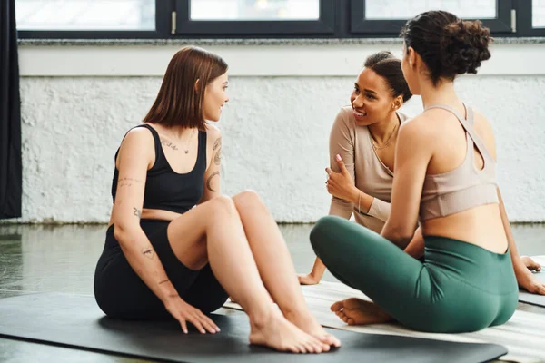 Young and positive african american woman in sportswear sitting on yoga mat and talking to tattooed girlfriend during yoga class in gym, friendship, harmony and mental health concept — Stock Photo