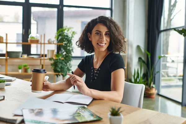Optimistic multiracial motivation coach holding takeaway drink in paper cup and smiling at camera near notebooks and magazines at workplace in interest club, social activism and dedication concept — Stock Photo