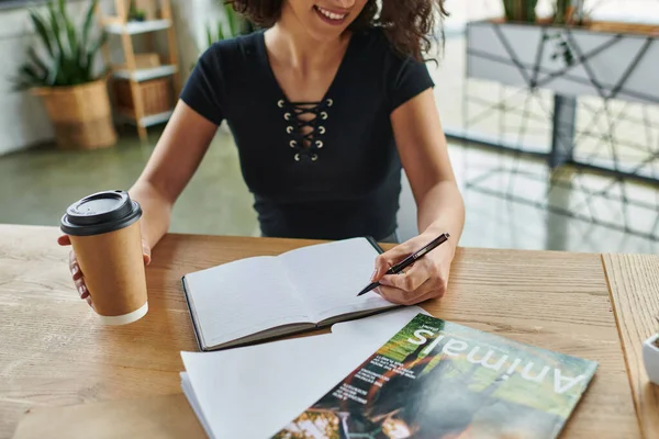 Personal growth, self-education, cropped view of smiling multiracial motivation coach with coffee to go writing in notebook near magazine in women interest club, social activism concept — Stock Photo