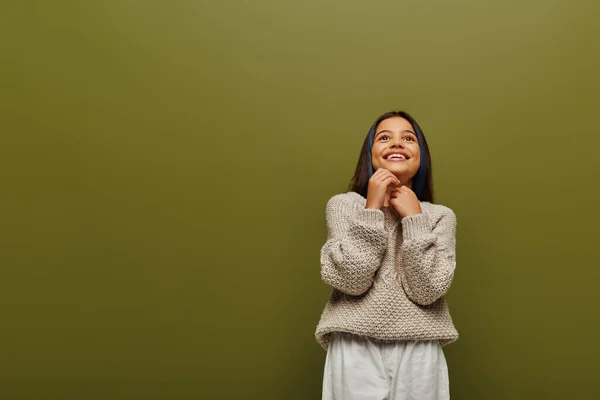 Мечтательная и улыбающаяся девочка-подросток с крашеными волосами в стильном трикотажном свитере смотрит в сторону, стоя и позируя изолированно на зеленой, современной моде для детского концепта — стоковое фото