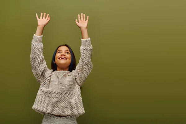 Morena positiva preadolescente niño con el pelo teñido en suéter de punto moderno levantando las manos y mirando hacia otro lado mientras está de pie aislado en verde, moda contemporánea para el concepto preadolescente - foto de stock