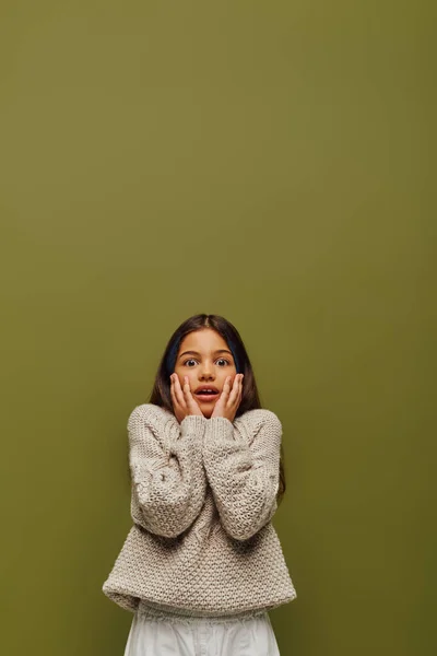 Shocked brunette preteen girl with dyed hair in casual and stylish knitted sweater touching cheeks and looking at camera isolated on green, contemporary fashion for preteen concept — Stock Photo