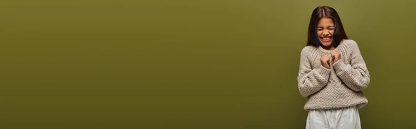 Muchacha preadolescente asustada en suéter de punto moderno y ropa casual cerrando los ojos mientras está de pie y posando aislado en verde, pancarta con espacio de copia, moda contemporánea para el concepto preadolescente - foto de stock