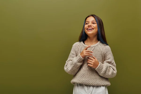 Счастливая и модная брюнетка-подросток с крашеными прядями волос в современном трикотажном свитере и отводя взгляд, стоя изолированно на зеленой, современной моде для детского концепта — стоковое фото