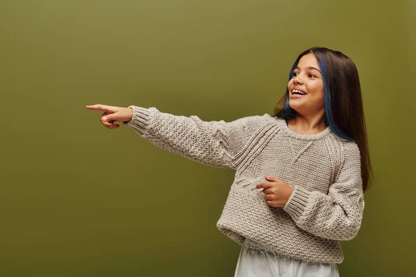 Cheerful and trendy preteen girl with dyed stands of hair wearing modern knitted sweater and pointing with finger isolated on green, contemporary fashion for preteen concept — Stock Photo