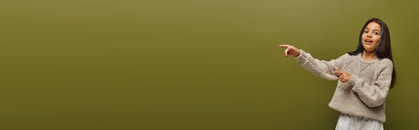 Muchacha preadolescente emocionada y de moda con mechones teñidos de pelo que usan suéter de punto y mirando a la cámara mientras posan con los dedos aislados en verde, bandera, moda contemporánea para el concepto preadolescente - foto de stock