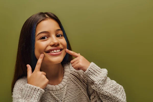 Portrait of cheerful preteen kid with dyed hair wearing modern knitted sweater and pointing with fingers at mouth while standing isolated on green, fashion-forward preteen with sense of style — Stock Photo