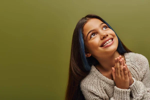 Portrait of smiling brunette preteen girl with dyed hair wearing knitted sweater while doing praying hands gesture and standing isolated on green, fashion-forward preteen with sense of style — Stock Photo